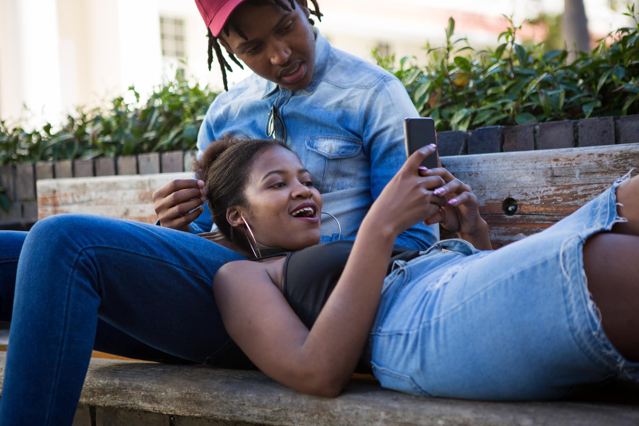 A young woman laughs whlie looking at her cellphone, lounging on a young man's lap. He is also looking at her phone screen