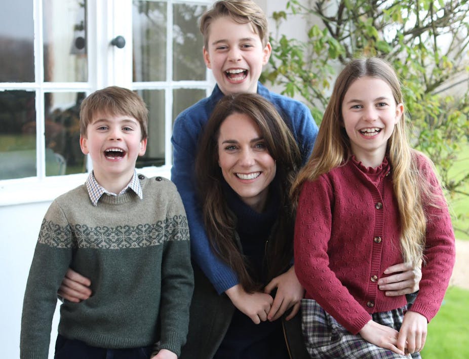 Family portrait of the Princess of Wales with her three children around her, all smiling and dressed casually. They are outdoors in front of a white glass paned window and a green leafed tree.