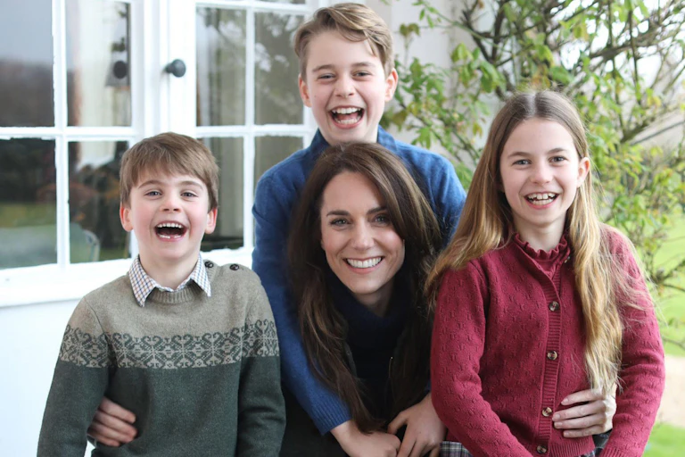 Family portrait of the Princess of Wales with her three children around her, all smiling and dressed casually. They are outdoors in front of a white glass paned window and a green leafed tree.