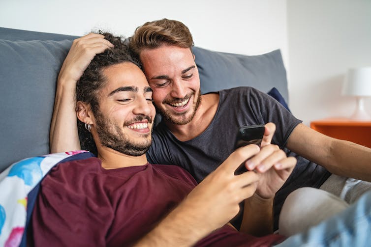 A gay couple lies in bed together and looks happily at a phone
