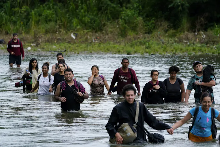 Uma fila de pessoas a atravessar a água com árvores da selva ao fundo.