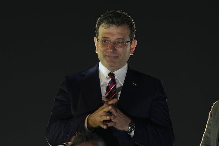 A man in a dark suit with his hands clasped.