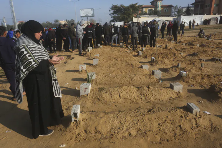 A woman wearing a hijab stands in front of a grave with her hands open