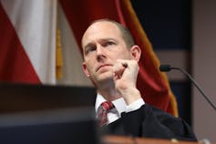 A middle aged white man wearing a black robe listens to testimony.