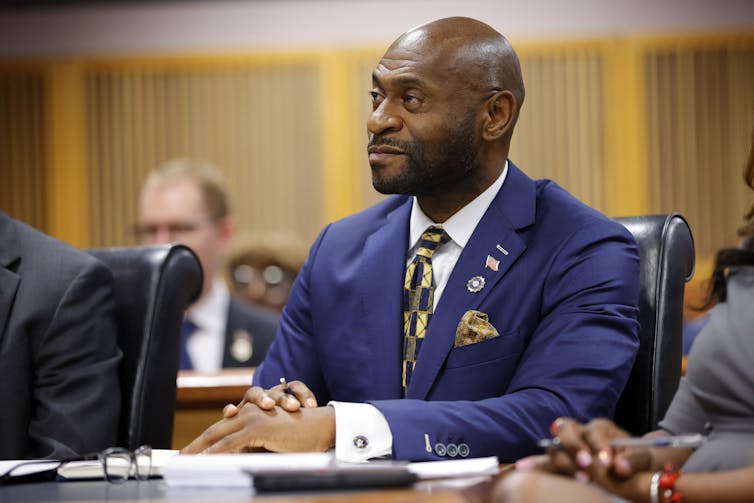 A Black man dressed in a dark suit sits at a table with his hands held together.