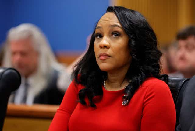 A Black woman wearing a red dress sits behind a desk as she listens to arguments from lawyers.