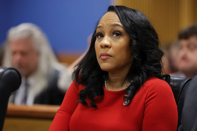 A Black woman wearing a red dress sits behind a desk as she listens to arguments from lawyers.