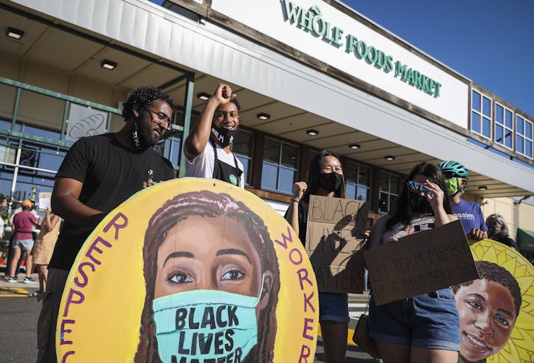 People stand in front of a Whole Foods with painted signs depicting a woman in a Black Lives Matter face mask and another one with a Black person's face without a mask.