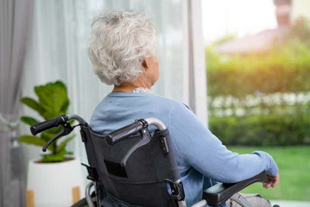 An elderly lady sitting in a wheelchair looks out the window.