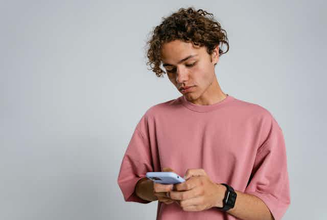 A teenage boy holds a phone against a blank background. 