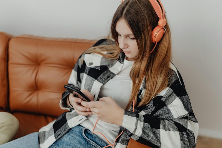 A teenager girl wearing headphones sits on a couch, looking at a phone.