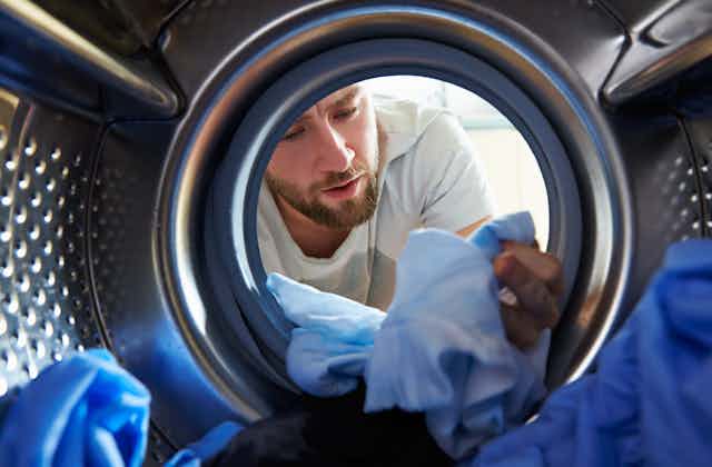 A view from a washing machine looking out at a bearded man inspecting his clothes in the drum.