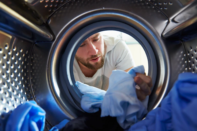 A view from a washing machine looking out at a bearded man inspecting his clothes in the drum.