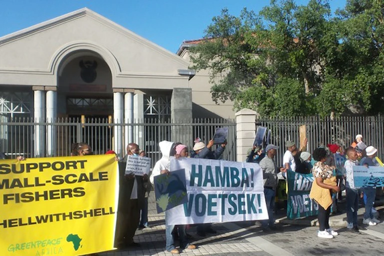 Protestors stand outside a courthouse carrying a yellow banner proclaiming 'support small scale fishers, To Hell with Shell' and another white banner proclaiming "Hamba! Voetsek!" which means get lost