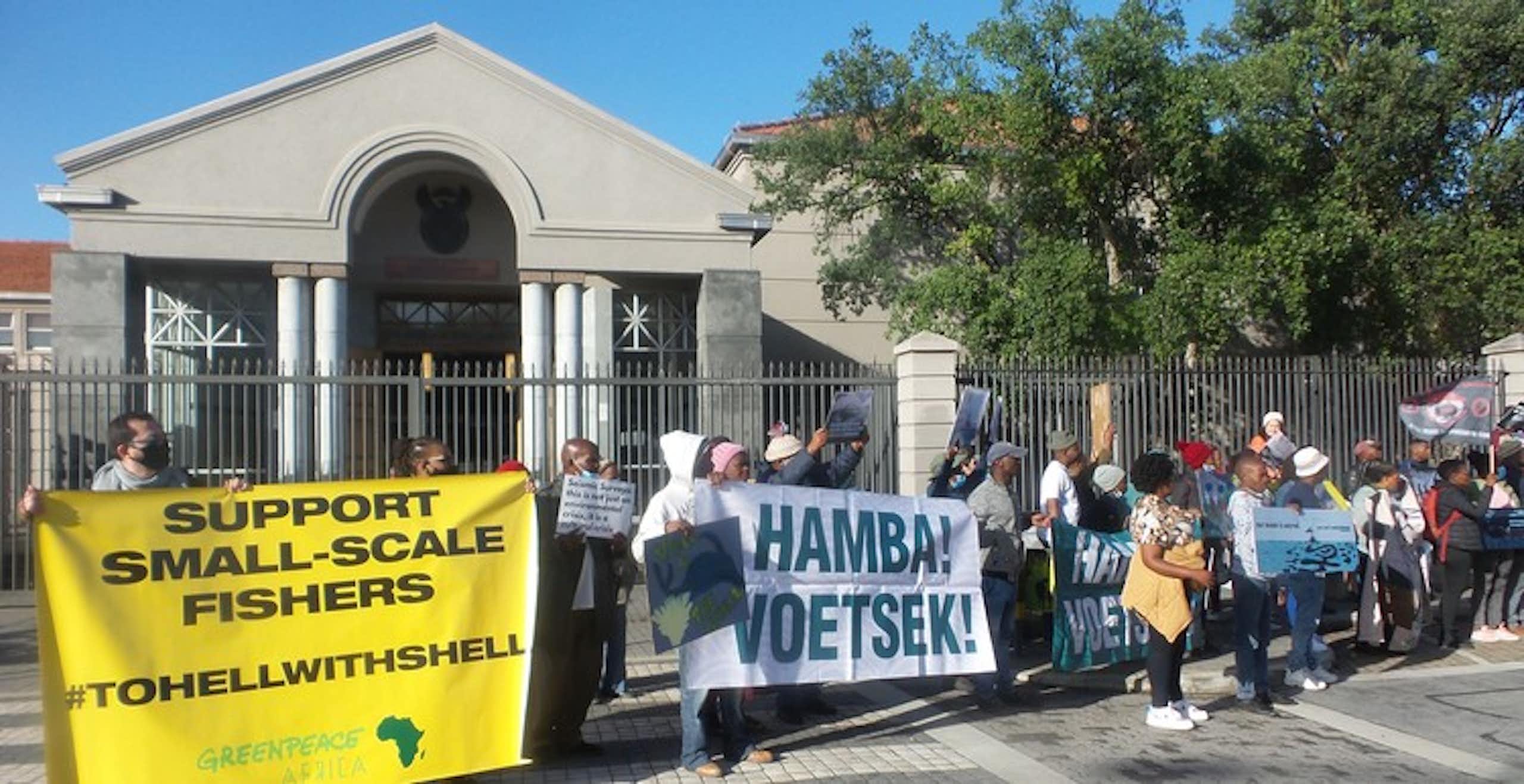 Protestors stand outside a courthouse carrying a yellow banner proclaiming 'support small scale fishers, To Hell with Shell' and another white banner proclaiming "Hamba! Voetsek!" which means get lost