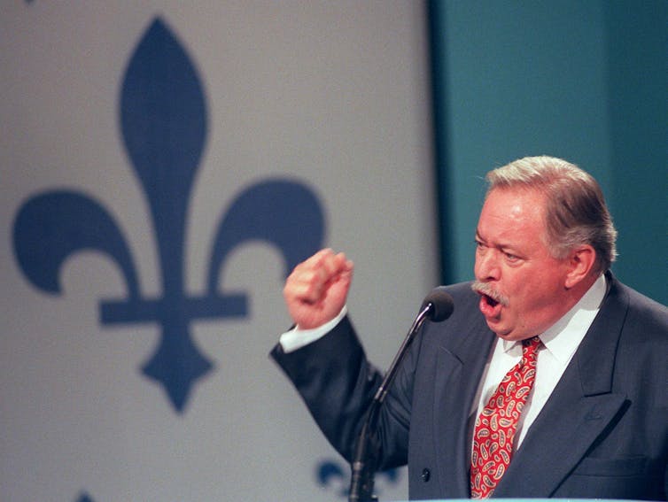 A man holds his fist up during a speech. The Quebec fleur-de-lis is in the background.