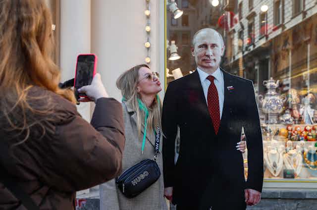 A woman takes a picture of her friend blowing a kiss at a cardboard figure of Russian president Vladimir Putin, March 2024