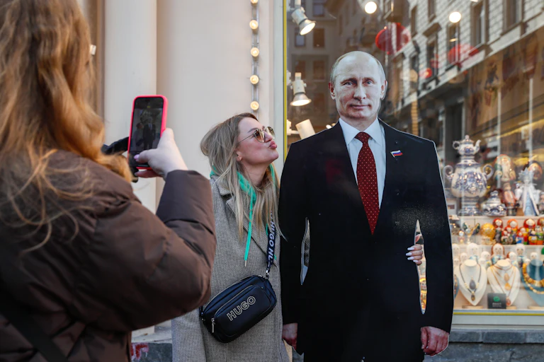 A woman takes a picture of her friend blowing a kiss at a cardboard figure of Russian president Vladimir Putin, March 2024