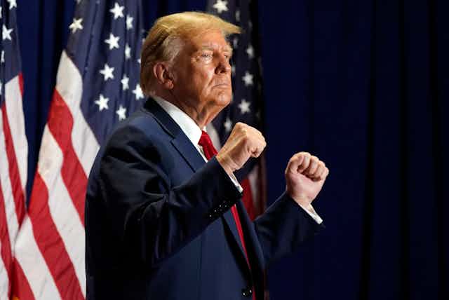 A man in a suit and tie clenches both fists while standing in front of American flags.