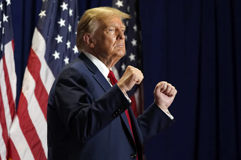 A man in a suit and tie clenches both fists while standing in front of American flags.