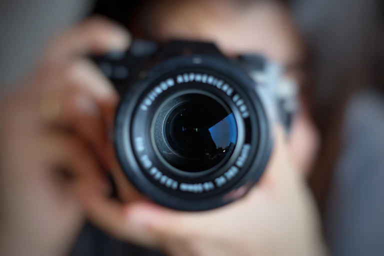 Close up of a photographer holding a camera to their eye, the lens is in focus, the photographer is out of focus