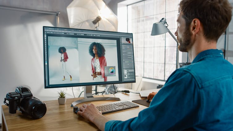 A man sits at a desktop computer and edits a photo of a model on an editing programme like Photoshop