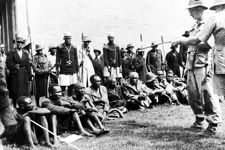 A black and white photo of two white men in uniform carrying guns standing in front of a group of black men seated on the ground. Other men are seen standing in the background.