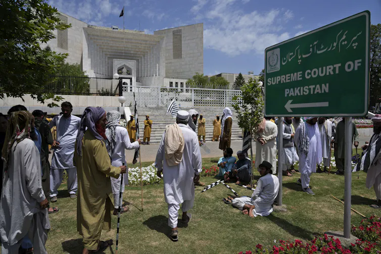 people walk passed a large white building. A sign pointing at it reads Supreme Court of Pakistan