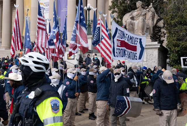 Several men in blue jackets holding American flags outside a building with Corinthian columns and a seated statue.