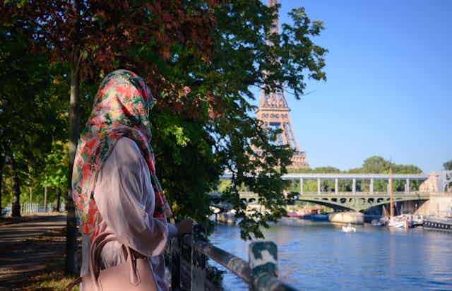 A woman in a floral hijab seen from behind standing by a river with the eiffel tower in the background