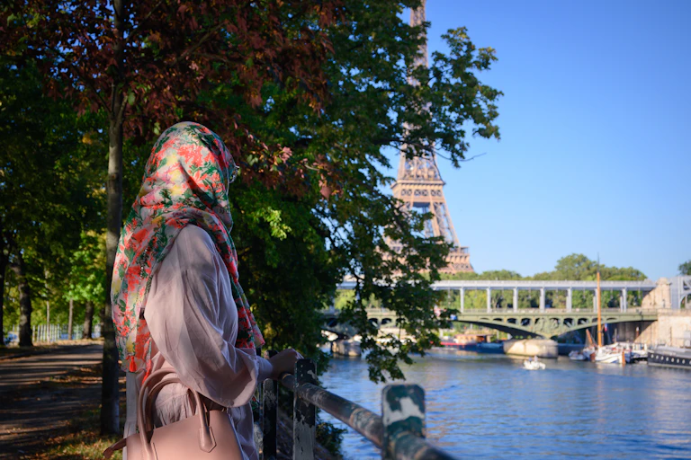 A woman in a floral hijab seen from behind standing by a river with the eiffel tower in the background