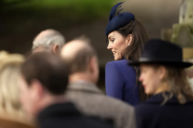 A woman in a blue hat and coat with long dark hair is seen from behind among family members.