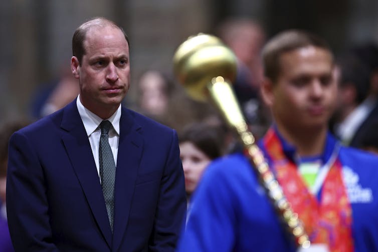 A balding man looks sombre walking behind a page dressed in a blue cloak carrying a golden rod.