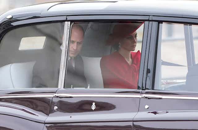 A balding man in a suit and a woman in a red hat and cloak sit in the back of a limousine looking glum..