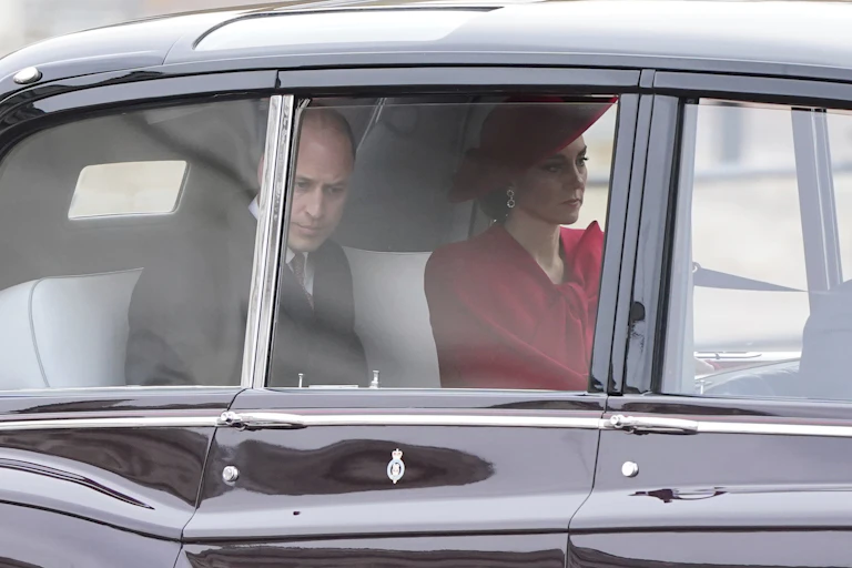 A balding man in a suit and a woman in a red hat and cloak sit in the back of a limousine looking glum..