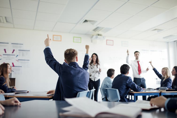 Students raise their hands in a classroom. Two teachers at the front look at students, selecting someone to respond.