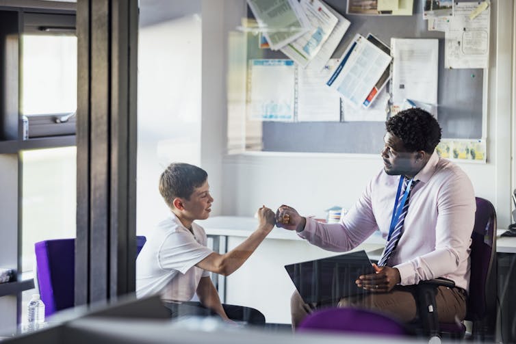 A male teacher fist bumps a male student. Both are seated in a classroom.