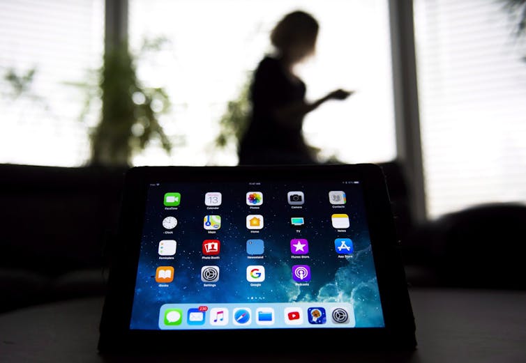 An iPad screen in the foreground and a person using a smartphone in silhouette in the background