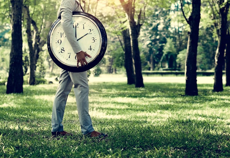 Man carrying large clock under his arm in park with trees