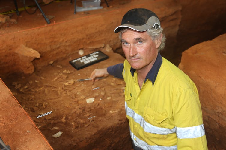 A man wearing a high vis jacket stands in a red rocky cave with archaeology tools in the background.