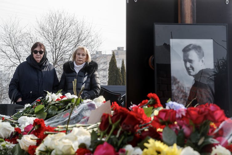 Photot of Alexey Navalny next to pile of flowers with two women in background