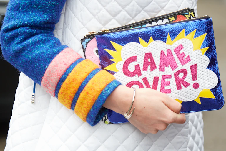 A woman's hand holding a clutch bag that says "GAME OVER!"