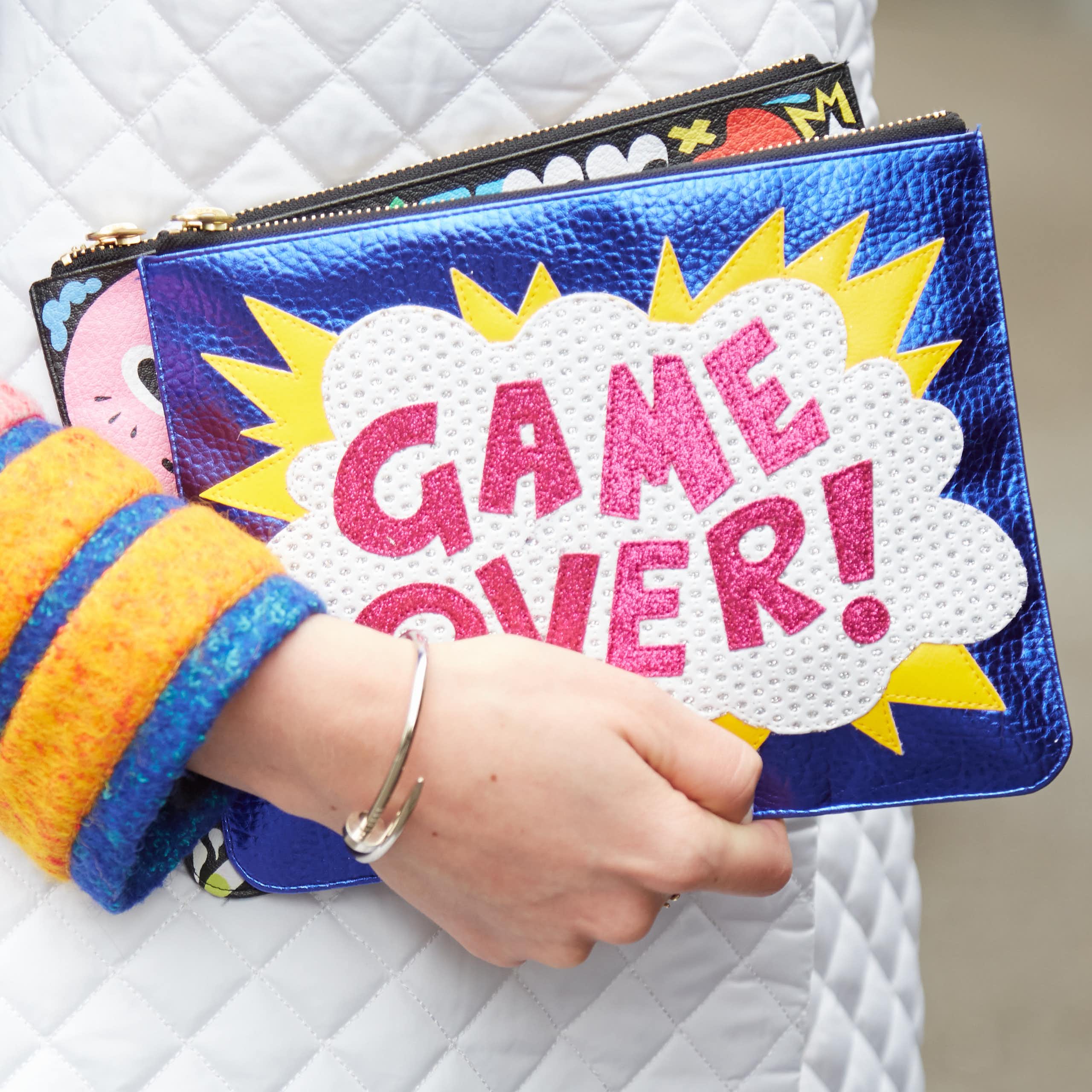 A woman's hand holding a clutch bag that says "GAME OVER!"