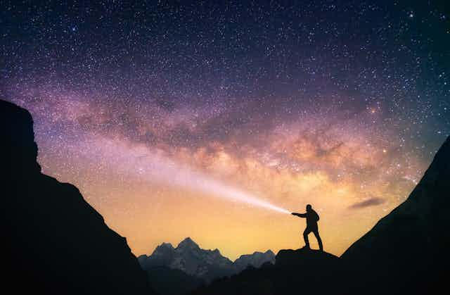 Silhouette of the man standing against the Milky Way in the mountains with a flashlight in his hands.