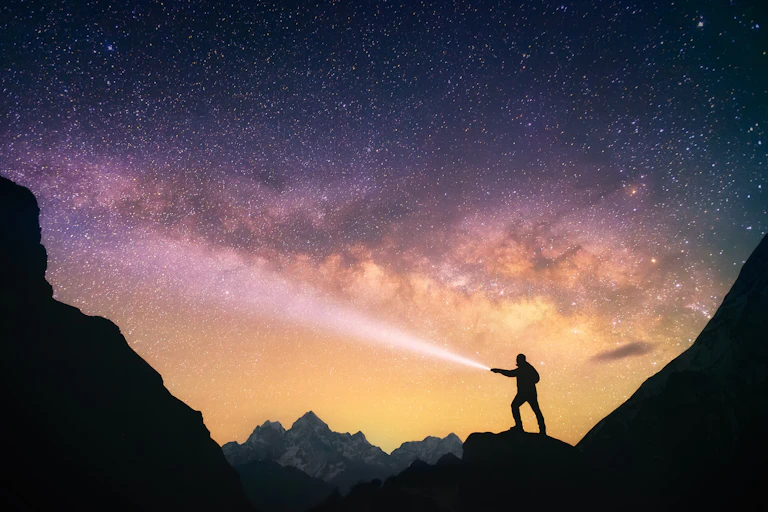 Silhouette of the man standing against the Milky Way in the mountains with a flashlight in his hands.