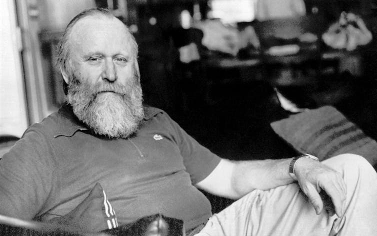 Black and white photo of beareded man sitting in a chair and posing for the camera.