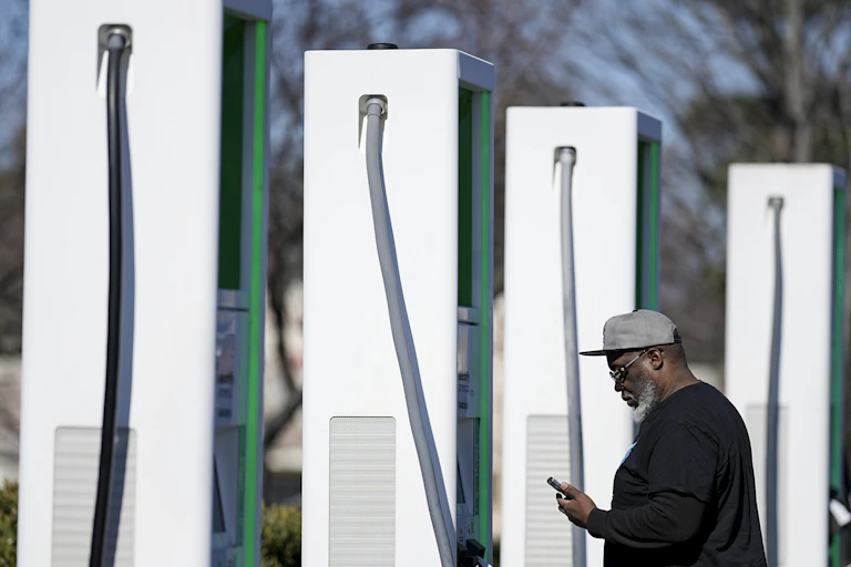 A man stands looking at his phone next to a row of EV chargers.