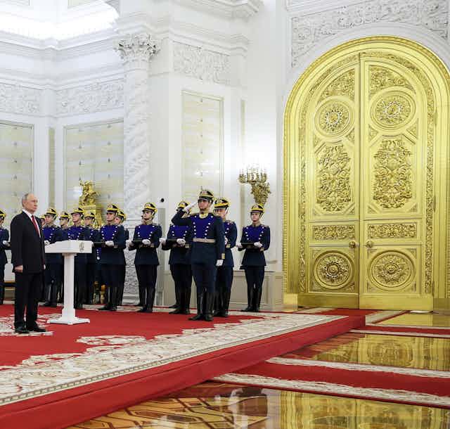 A man stands behind a podium flanked on each side by rows of soldiers and citizens. A massive golden door is beside him.