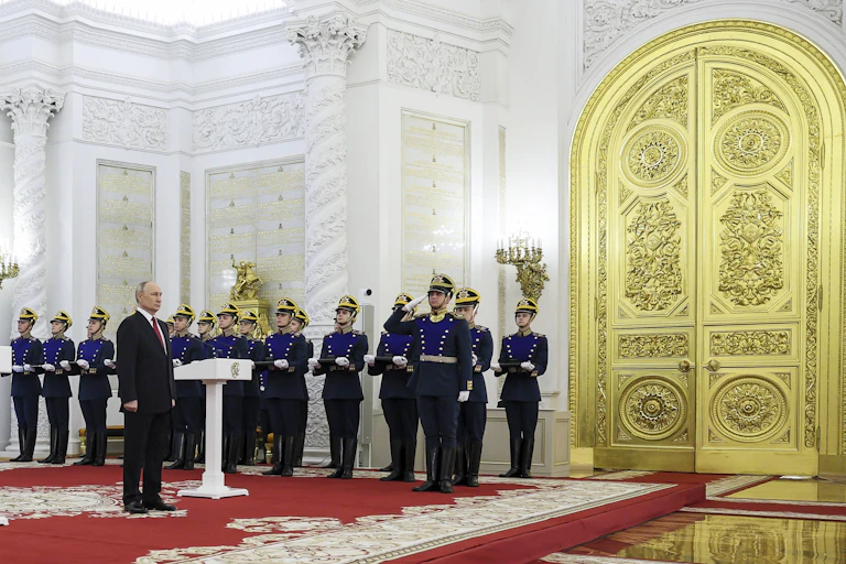 A man stands behind a podium flanked on each side by rows of soldiers and citizens. A massive golden door is beside him.