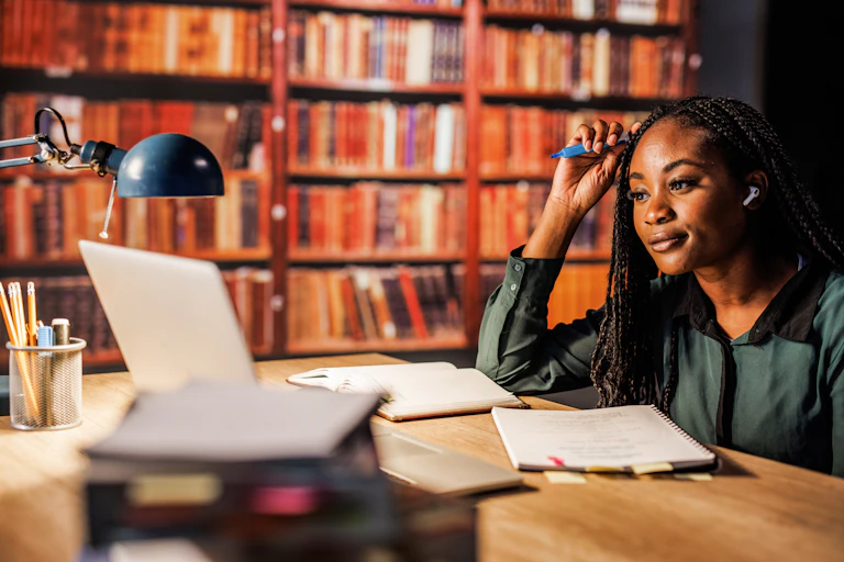A Black woman with braids sits at a table in a library.